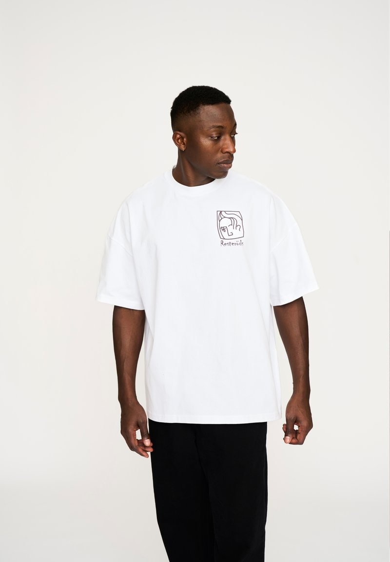 Hvid bomulds t-shirt med en afslappet pasform, prydet med et lilla grafisk design på venstre brystområde. Bæres med sorte bukser.