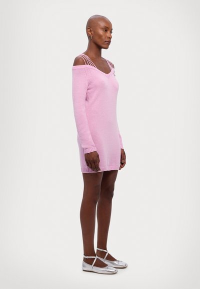 Robe tricotée rose avec un design décolleté, manches longues et poignets côtes. Le modèle porte des ballerines argentées avec des brides de cheville.