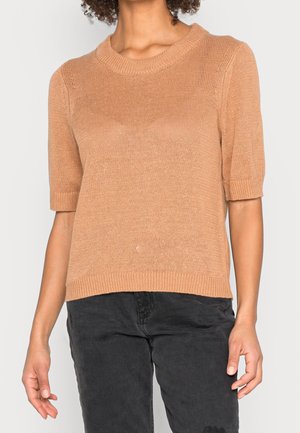 Femme portant un pull en tricot camel à manches courtes associé à un pantalon noir, debout devant un fond clair.
