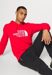 Sweatshirt com capuz vermelho, com impressão de logótipo branco, cordão e bolso canguru; combinado com calças pretas; textura suave e ajuste casual.