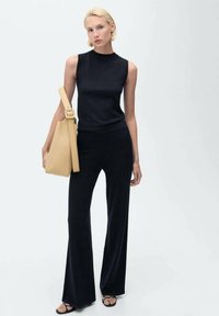 Čierny top bez rukávov s vysokým golierom, v kombinácii s čiernymi zvonovými nohavicami. Doplnok: béžová ramenná taška s okrúhlym uchom.