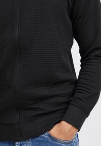 Sudadera con cremallera negra de textura acanalada, con puños y cinturilla ajustados. La tela parece suave y ligeramente elástica.