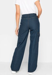 Siniset leveälahkeiset farkut, joissa on korkea vyötärö, sivuissa cargo-taskut ja kontrastiväriset langan ompeleet, yhdistettynä valkoiseen t-paitaan.