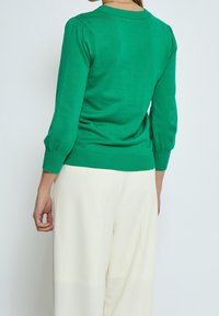 Grüner Pullover mit Rundhalsausschnitt und dreiviertellangen Puffärmeln, mit gerippten Bündchen und Saum, getragen über weißen Hosen.