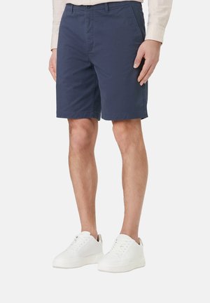 Hombre vistiendo pantalones cortos azul marino hasta la rodilla, zapatillas blancas y una camisa de manga larga beige claro contra un fondo liso.