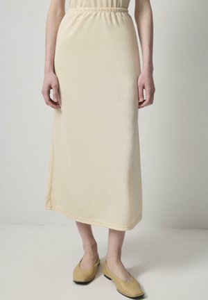 Dlhá béžová sukňa s elastickým pásom, vyrobená z hladkej, ľahkej tkaniny, dosahujúca do polovice lýtka. Kombinovaná s jednoduchými žltými balerínkami.