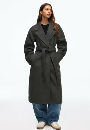 Femme portant un manteau en laine gris foncé ceinturé, un jean bleu large et des baskets blanches, debout avec les mains dans les poches du manteau.