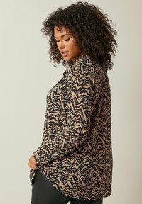 Camicia a maniche lunghe color sabbia con un motivo a zigzag nero, dotata di colletto, chiusura con bottoni e orlo curvo. Tessuto morbido con una sensazione strutturata.