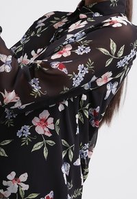 Blouse noire à fleurs avec un col, des manches longues et un tissu transparent orné de fleurs rouges et bleues ainsi que de papillons.