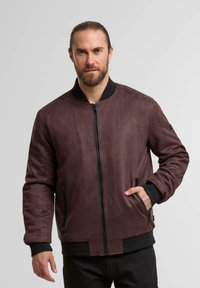 Jachetă bomber din suede maro, cu un guler ondulat negru, închidere cu fermoar și buzunare laterale. Textură moale, cu o culoare uniformă și un design casual.