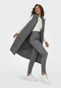 Gilet oversize gris en duvet avec capuche, présentant un design matelassé, associé à un pantalon gris fuselé et des baskets blanches. Texture lisse, coupe décontractée.