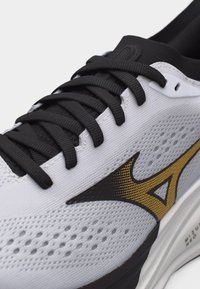 Λευκό αθλητικό παπούτσι με μαύρα κορδόνια, μαύρο και χρυσό λογότυπο και διαπνέον επάνω μέρος με τρυπητό σχέδιο, κοντινή λήψη στο πλάι και την περιοχή των δαχτύλων.