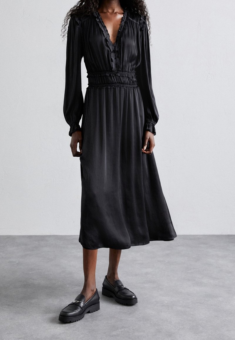 Femme portant une robe midi noire à manches longues avec des détails en volants au niveau de l'encolure et de la taille, associée à des mocassins noirs, debout sur un sol gris.