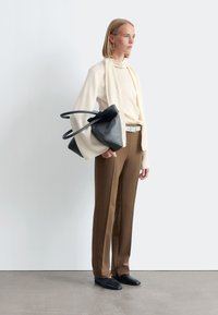 Black leather handbag with a unique shape and double handles; cream high-neck top; brown trousers; black slip-on shoes; minimalistic design.