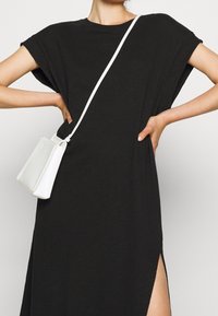 Černé oversize tričko s krátkými rukávy a postranními rozparky, doplněné malou bílou texturovanou kabelkou přes rameno a tenkým bílým popruhem.