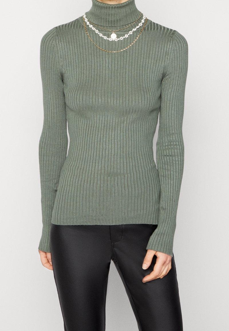 Femme portant un pull col roulé ajusté olive vert côtelé et un pantalon en cuir noir, accessoirisée avec des colliers superposés en or et à motifs floraux blancs.