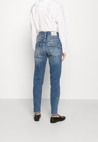 Modré džíny s vysokým pasem a úzkým střihem, s kapsami na zadní straně a jemným vyblednutím. Stylizované s bílou košilí a černými mokasíny.