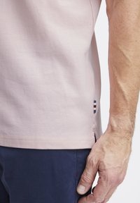 Világos rózsaszín pólóing texturált anyagból, oldalsó hasítékkal az alján és egy kis többcsíkos díszítéssel a bal oldalon.