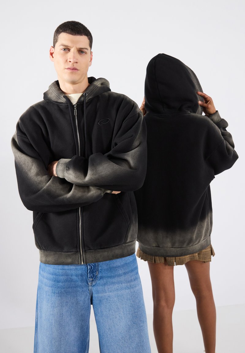 Felpa nera con zip e sfumatura grigia, dotata di tasca frontale e cappuccio con cordini. Modelli presentati frontalmente e posteriormente, indossando jeans blu.