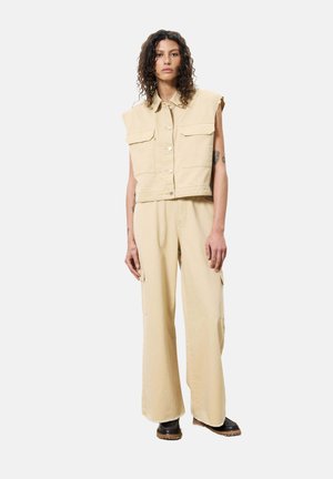 Beige denim mouwloze jas met twee borstzakken, gecombineerd met beige wijde broekspijpen met zijzakken en een licht rafelige zoom.
