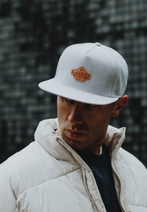 Grijze platte pet met een tan leren label "BLACKSKIES," gedragen met een witte pufferjas en een donker tshirt. Textuurstoffen zijn zichtbaar.