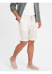 Shorts en lin léger couleur blanc cassé avec une taille élastique, des poches latérales et un ourlet roulé. Associés à des chaussures à enfiler beiges.