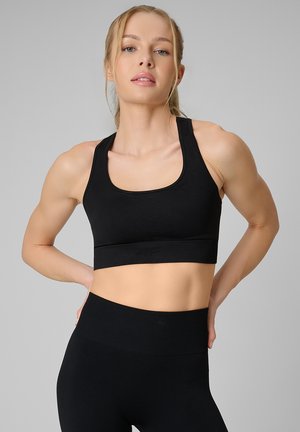 Kobieta z blond włosami w czarnym sportowym biustonoszu i legginsach stoi pewnie na tle jednolitego szarego tła, z rękami na biodrach.