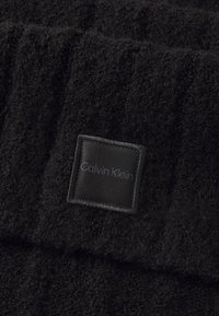 Asciugamano nero in morbido tessuto con superficie testurizzata. Presenta un'etichetta quadrata in tessuto nero con la scritta "Calvin Klein" impressa in grigio su un lato.