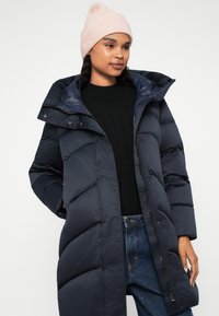 Abrigo de puff azul oscuro con capucha, con un diseño acolchado, cremallera frontal y botones a presión. Combinado con un suéter negro y jeans de mezclilla.