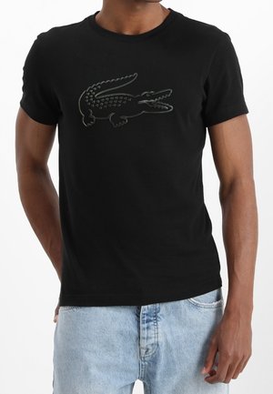 Homme portant un t-shirt noir avec un grand logo crocodile en relief sur la poitrine, associé à un jean bleu clair.