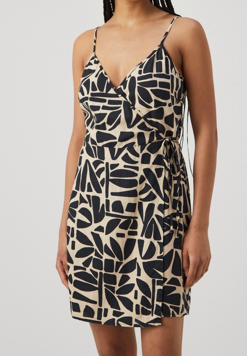 Femme portant une robe portefeuille beige sans manches avec des motifs géométriques abstraits noirs et une ceinture nouée sur le côté à la taille.
