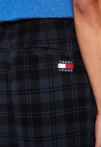 Czarne spodnie z tkaniny w kratę mają tylną kieszeń z prostokątną metką logo Tommy Jeans w granatowym, czerwonym i białym kolorze.
