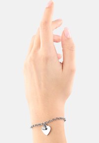 La pulsera de cadena plateada presenta un charm en forma de corazón con un acento de gema clara. Está llevada en una mano de piel clara sobre un fondo neutro.