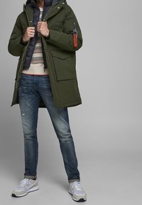 Olive green parka with a gray insulated inner layer, ribbed cuffs, front pockets, and an orange tag; paired with blue jeans and white sneakers.