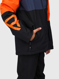Persona che indossa una giacca nera e arancione con una mano parzialmente dentro una tasca zip sul lato destro, su uno sfondo semplice.