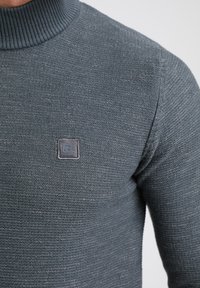 Harmaa, teksturoitu poolapaita, jossa on ribattu kaulus ja pieni logopatch rinnan kohdalla, korostaa kudontakuviota.