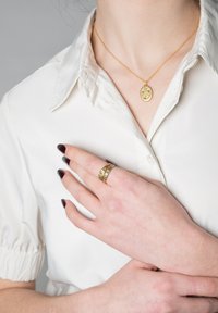Osoba s tmavým lakom na nechtoch nosí zlatý kvetinový prsteň a zodpovedajúci oválny prívesok na retiazke cez bielu košeľu s golierom.