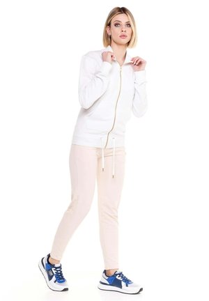 Une femme porte une veste blanche zippée, un pantalon clair et des baskets bleues et blanches sur un fond blanc uni.