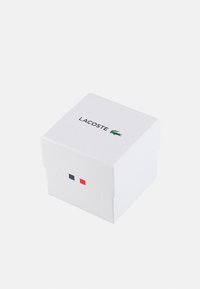 Valge ruudukujuline karp kaanega, millel on must "LACOSTE" logo, roheline krokodilli emblem ning punased ja sinised ristkülikukujulised aktsendid ühel küljel.