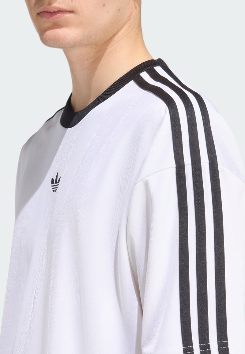 Camisa atlética blanca con cuello acanalado negro, que presenta tres franjas verticales negras en cada hombro y un logo bordado en el pecho.