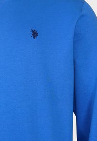 Blå långärmad skjorta i mjukt tyg. Har en mörk broderad logotyp på vänster bröst. Slät textur med en vanlig passform.