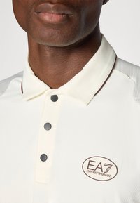 Λευκό πόλο μπλουζάκι με υφασμάτινη υφή, με καφέ περίγραμμα στον γιακά, δύο σκούρα κουμπιά και ένα οβάλ λογότυπο στο αριστερό μέρος.