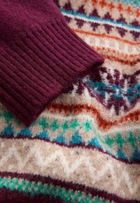 Bordeaux gebreide mouw met ribbelstructuur die een patroonstof overlayt met crèmekleurige, teal, oranje en paarse geometrische ontwerpen.