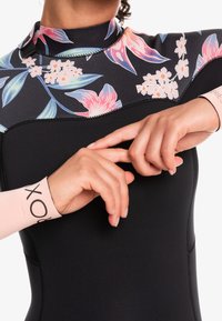 Black wetsuit with vibrant floral pattern on the neckline. Features long sleeves with light pink cuffs inscribed with "ROXY." Smooth texture.