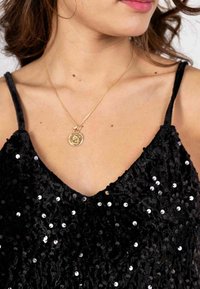 Top en sequins noirs avec un décolleté en V et des fines bretelles. Un collier pendentif en or est superposé au niveau du décolleté.