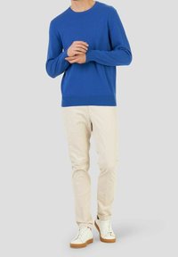 Man som bär en ljusblå långärmad tröja, beige byxor och vita sneakers med bruna sulor, stående mot en enfärgad bakgrund.