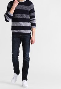 Homme portant un pull rayé bleu marine et gris, un jean foncé et des baskets blanches, marchant devant un fond blanc.
