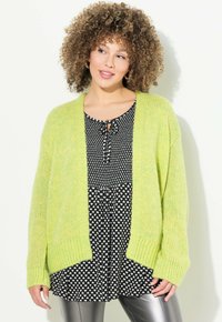 Gilet en tricot vert clair avec des poignets côtelés, porté sur une robe noire et blanche à pois avec un décolleté à nouer et une coupe ample.