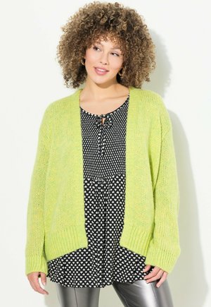 Gilet en tricot vert clair avec des poignets côtelés, porté sur une robe noire et blanche à pois avec un décolleté à nouer et une coupe ample.