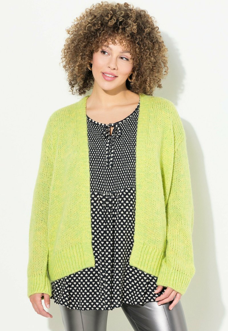 Gilet en tricot vert clair avec des poignets côtelés, porté sur une robe noire et blanche à pois avec un décolleté à nouer et une coupe ample.